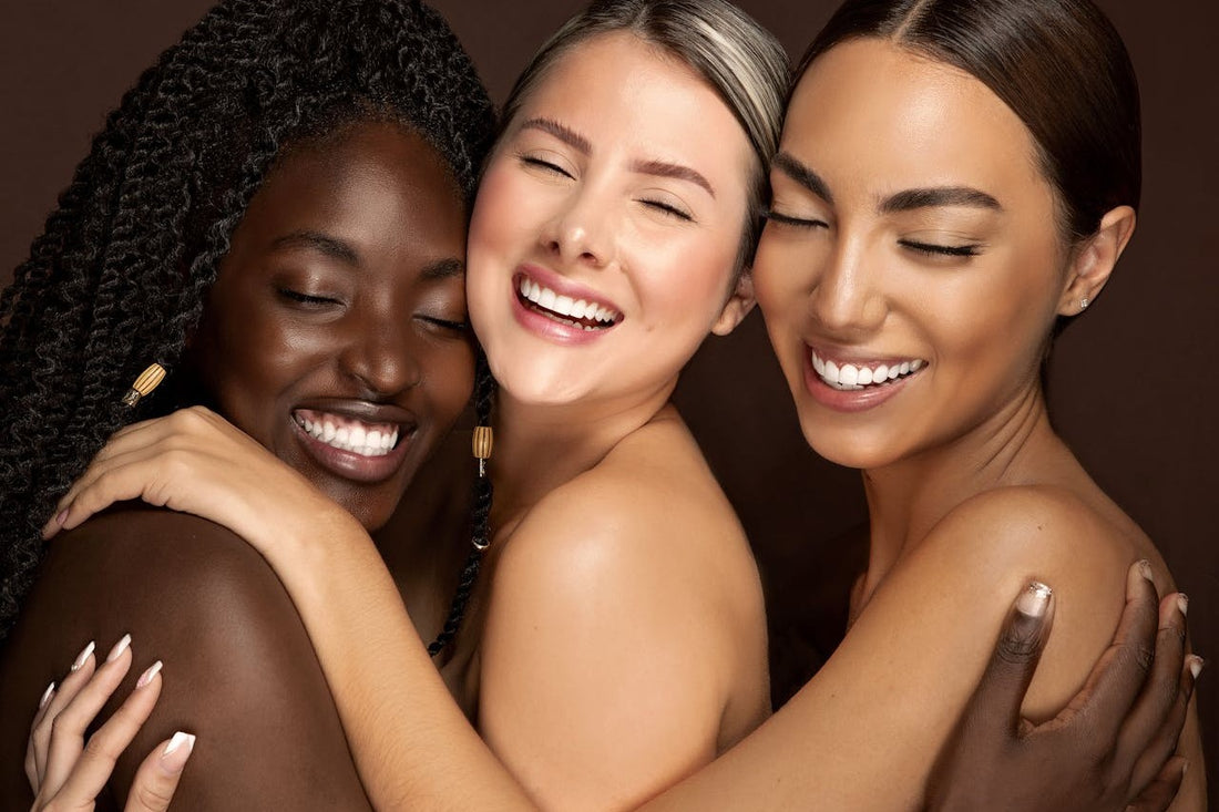 Three women with different skin tones embracing each other against a brown background