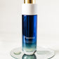 A bottle of LILIXIR Flawless Oil-to-Milk face cleanser on a clear glass coaster against a white background. The bottle is blue with a white cap and a gold accent ring.
