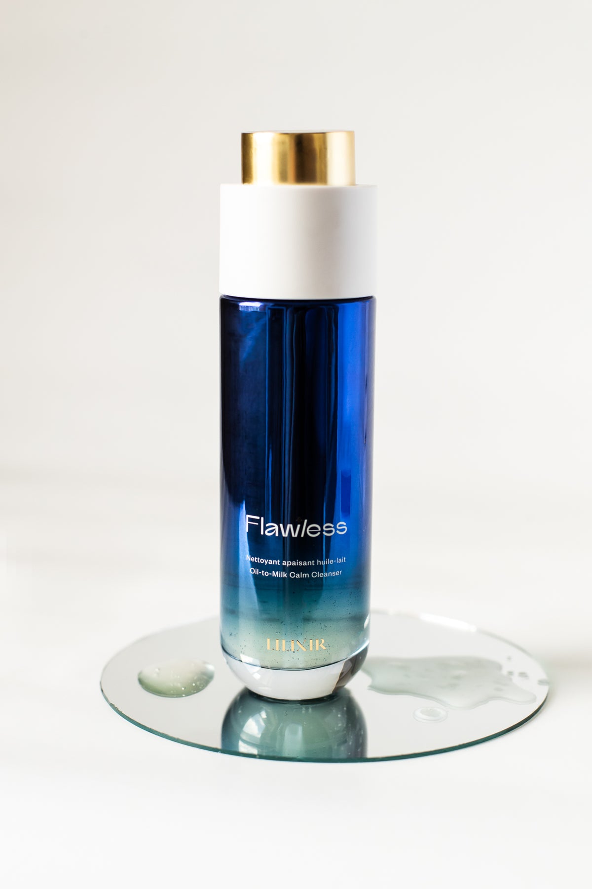 A bottle of LILIXIR Flawless Oil-to-Milk face cleanser on a clear glass coaster against a white background. The bottle is blue with a white cap and a gold accent ring.