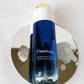 A bottle of LILIXIR Flawless Oil-to-Milk Cleanser with a blue gradient body, white cap, and gold cap accent, displayed on a reflective gold circular stand against a white background.