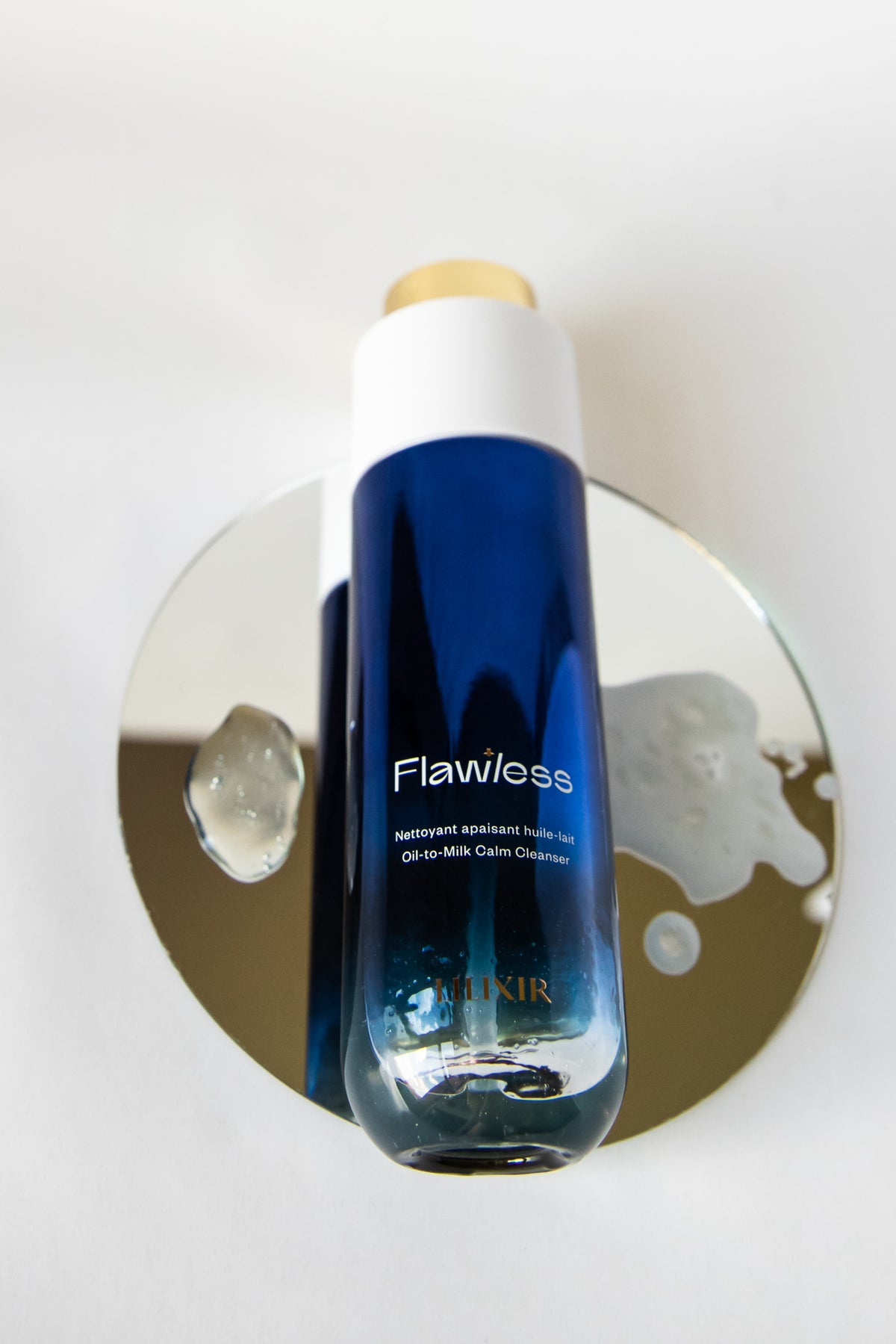 A bottle of LILIXIR Flawless Oil-to-Milk Cleanser with a blue gradient body, white cap, and gold cap accent, displayed on a reflective gold circular stand against a white background.