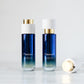Two bottles of LILIXIR Flawless Oil-To-Milk Calm Cleanser with a blue gradient body, white cap, and gold accent, displayed on a clear glass coaster against a white background.