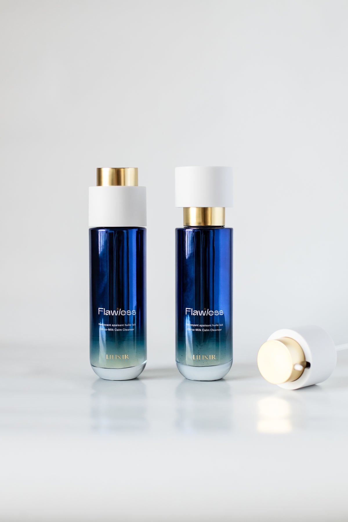 Two bottles of LILIXIR Flawless Oil-To-Milk Calm Cleanser with a blue gradient body, white cap, and gold accent, displayed on a clear glass coaster against a white background.