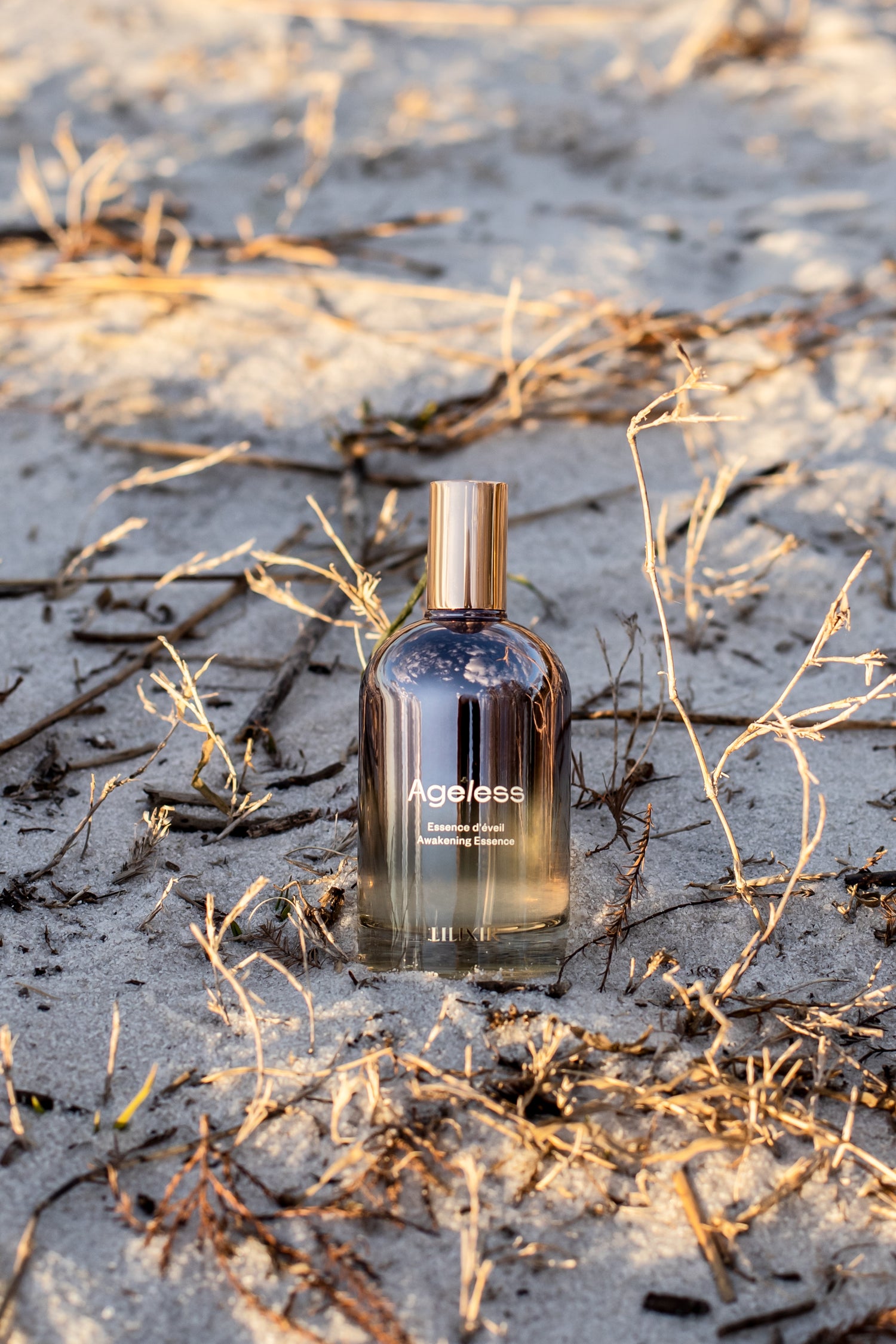 LILIXIR Ageless Awakening Essence  bottle on a sandy beach with dry grass.