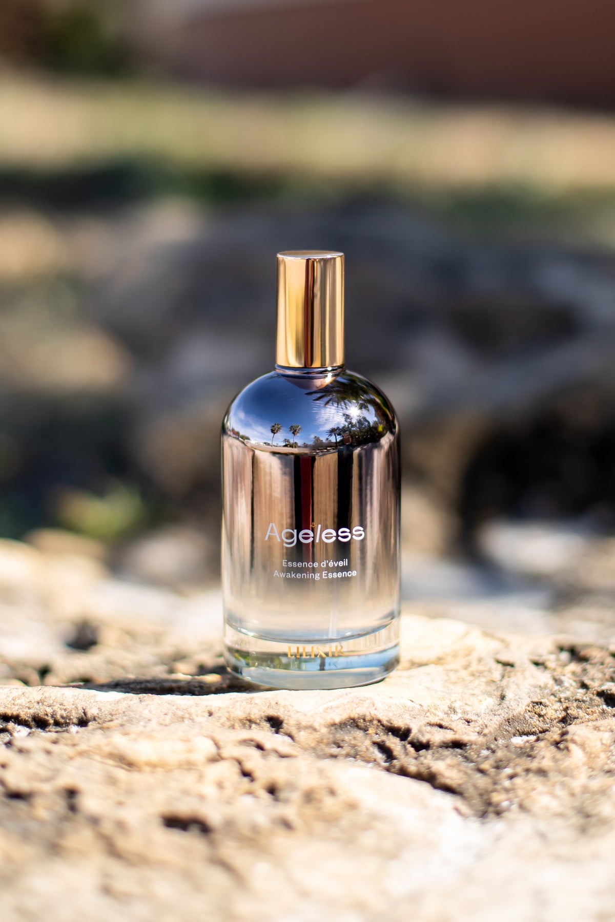 Clear bottle with gold cap labeled 'LILIXIR Ageless Awakening Essence' on a rock with blurred natural background