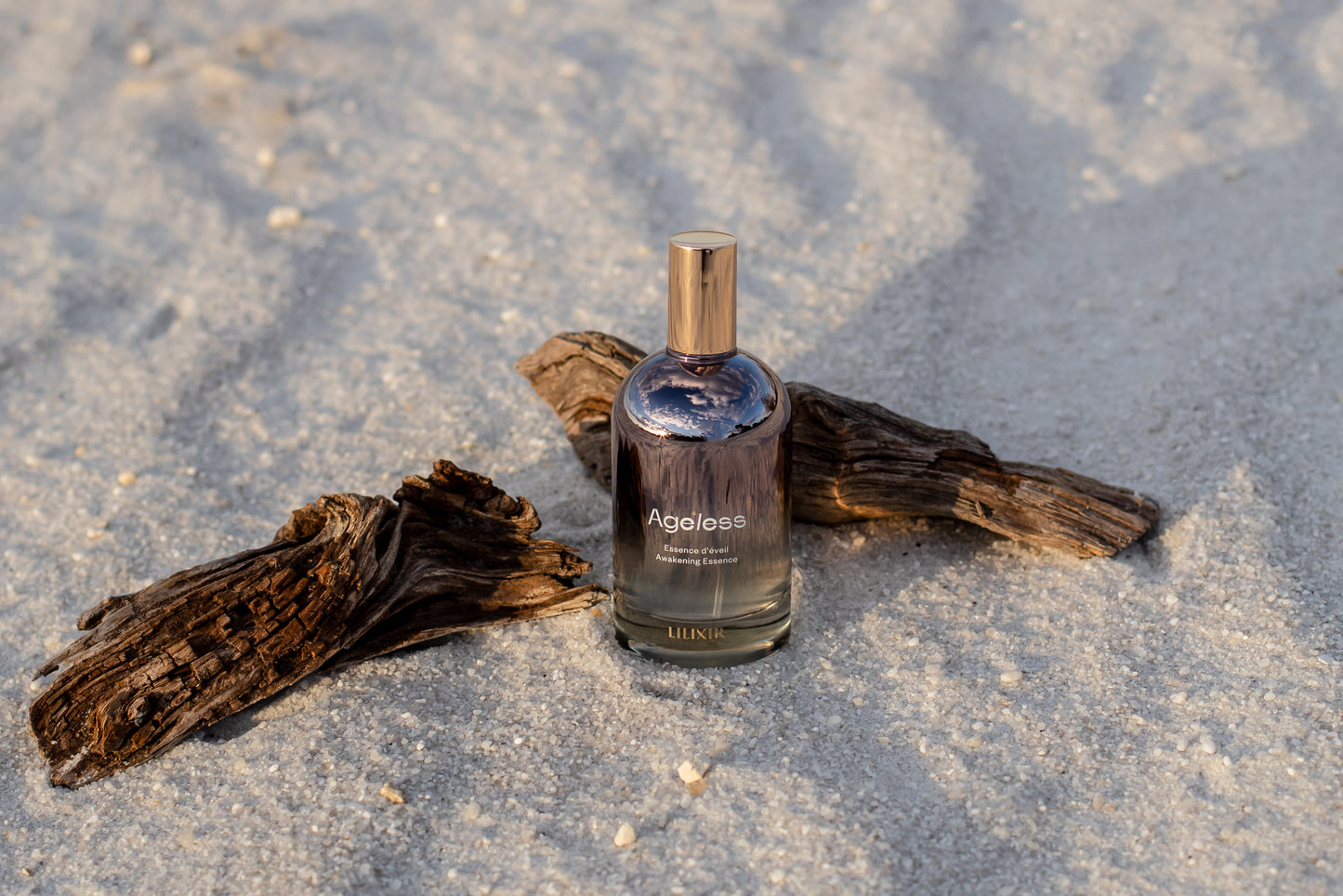 Bottle of 'LILIXIR Ageless Awakening Essence'  on a sandy surface with driftwood
