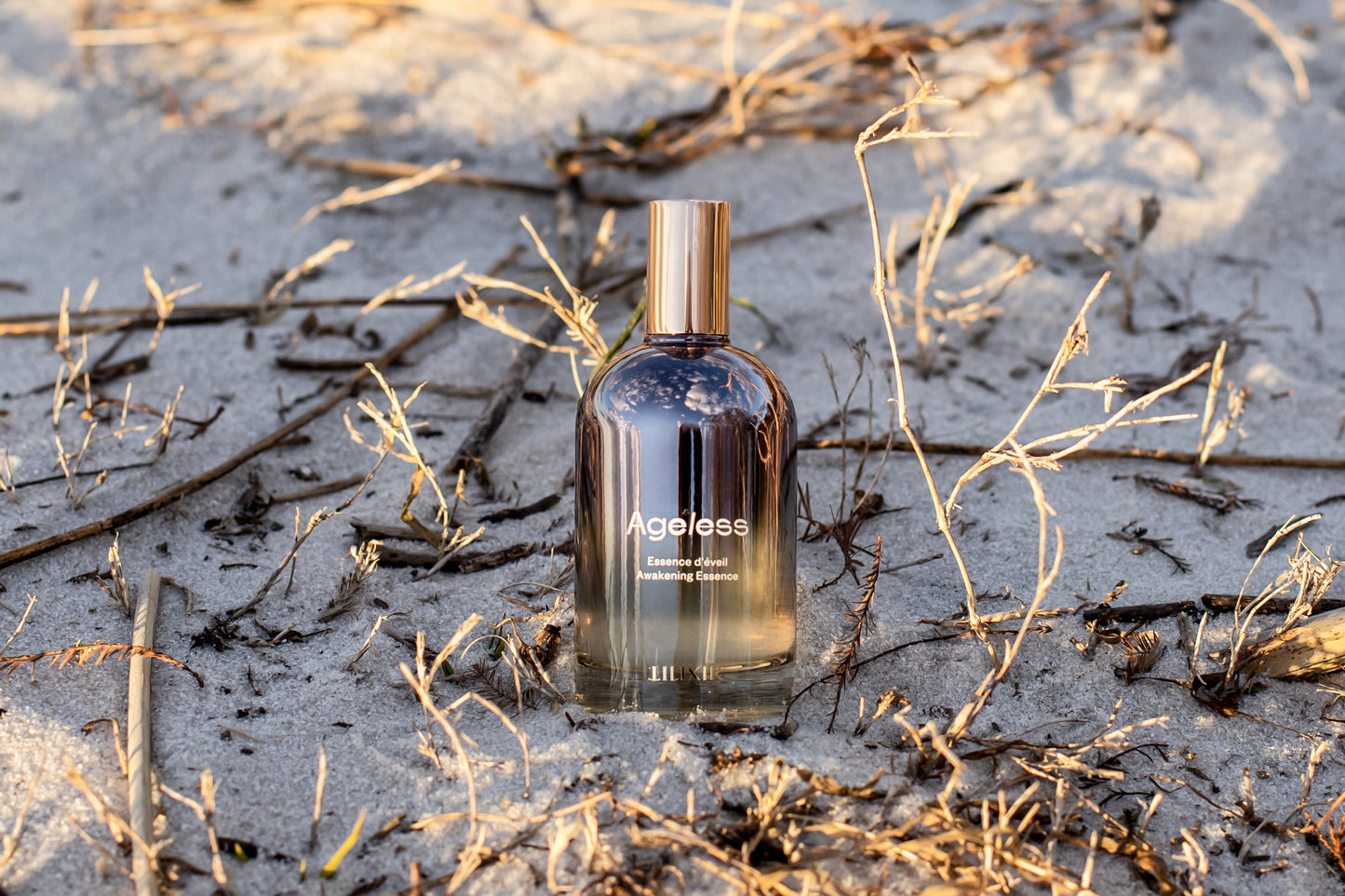 LILIXIR Ageless Awakening Essence' bottle on a sandy beach with dry grass