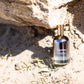 Bottle of 'LILIXIR Ageless Awakening Essence' on a sandy surface with rocks in the background