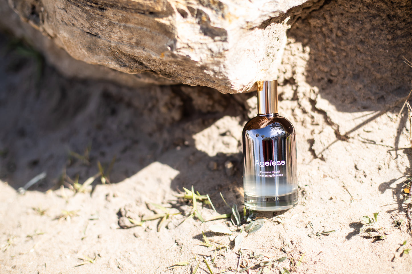 Bottle of 'LILIXIR Ageless Awakening Essence' on a sandy surface with rocks in the background