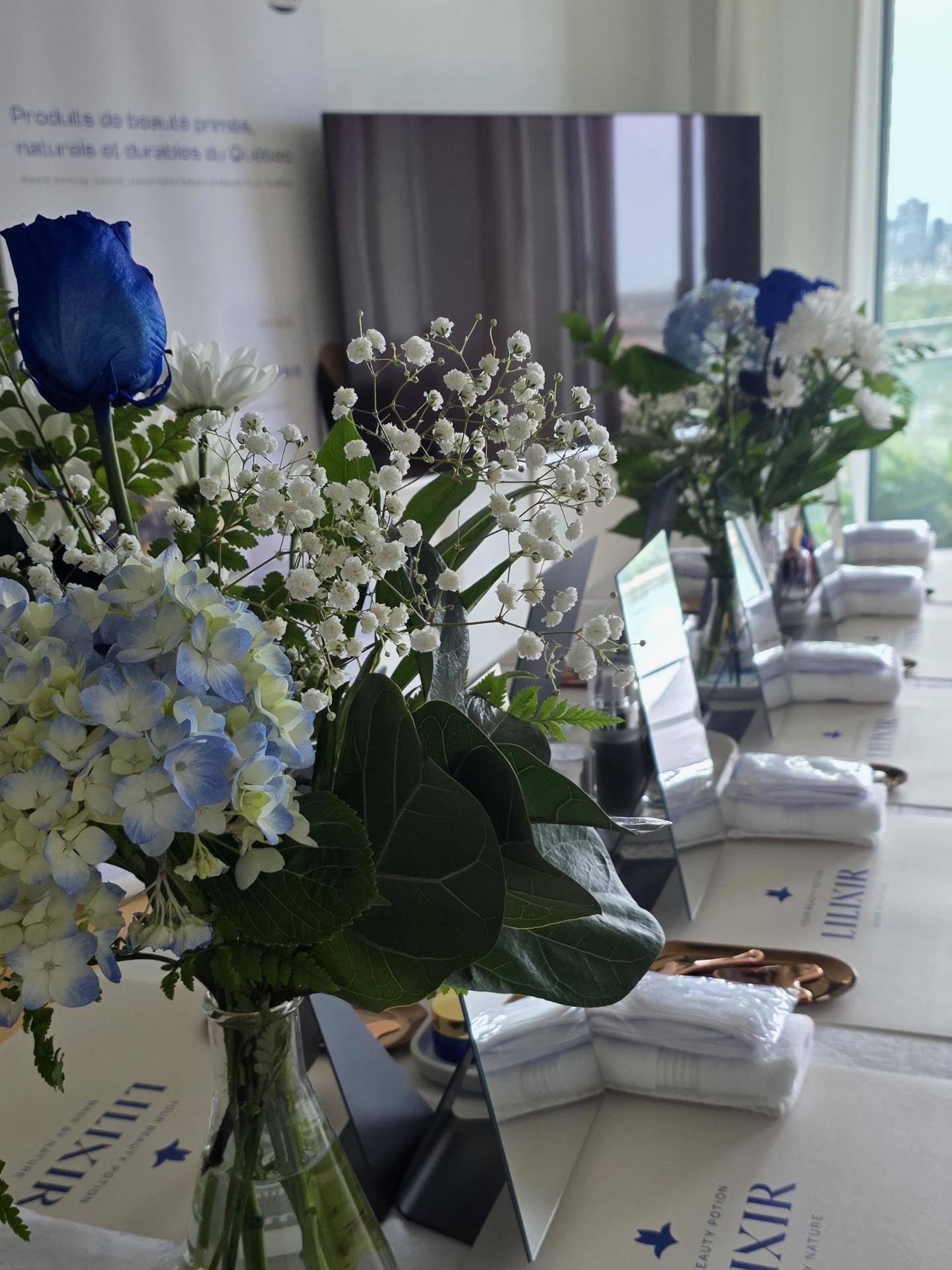 A styled table setting with floral arrangements in blue, white, and light blue hydrangeas and roses, alongside folded white towels and branded AXJR packaging on a light-colored surface.
