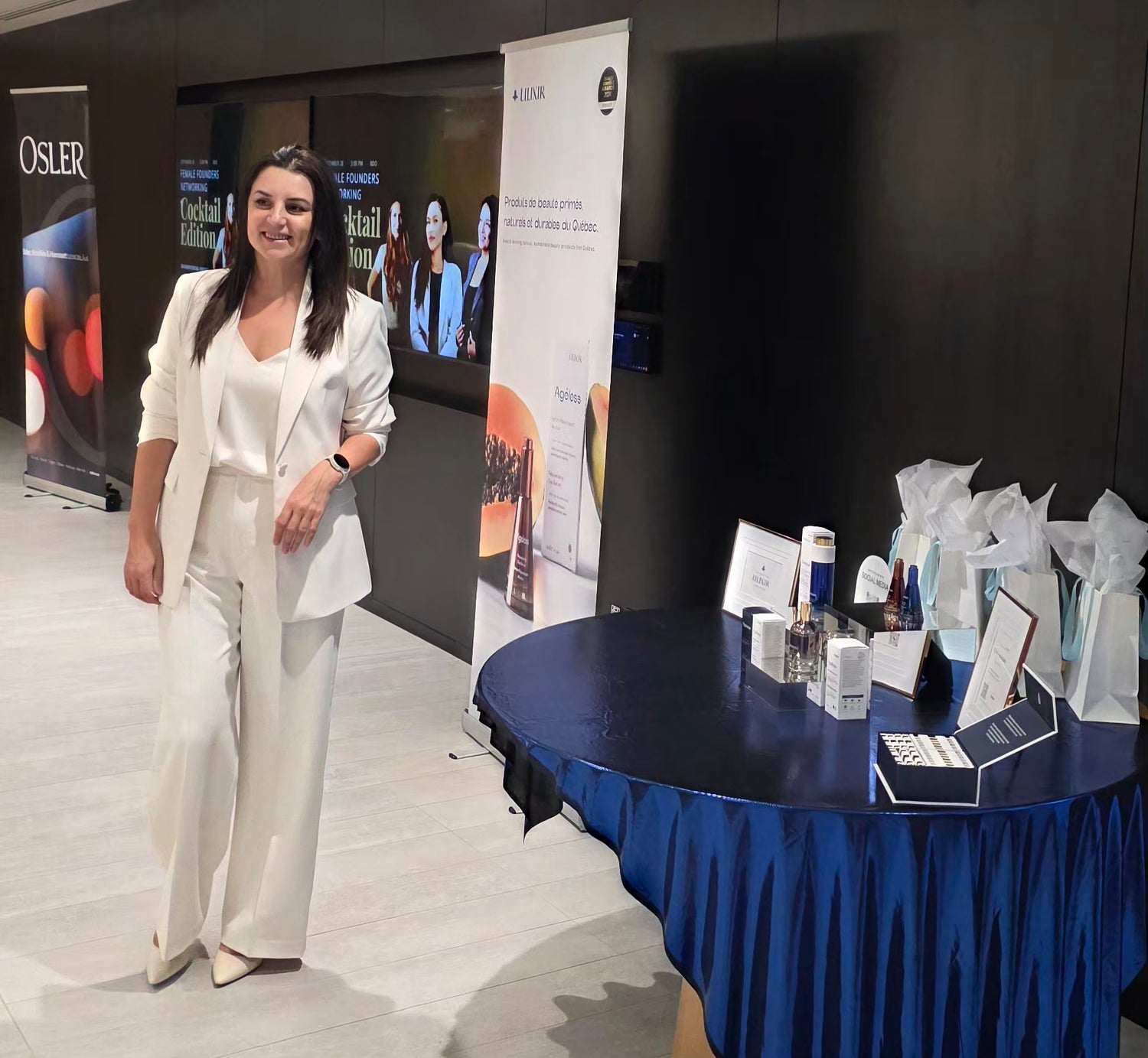 A woman in a white satin-like blazer, Lilia Ivanov the Founder of LILIXIR, and wide-leg trousers stands next to a round table with a blue drape and lilixir products on  the table in an indoor event space. A tall vertical banner and other event materials are visible behind her. 